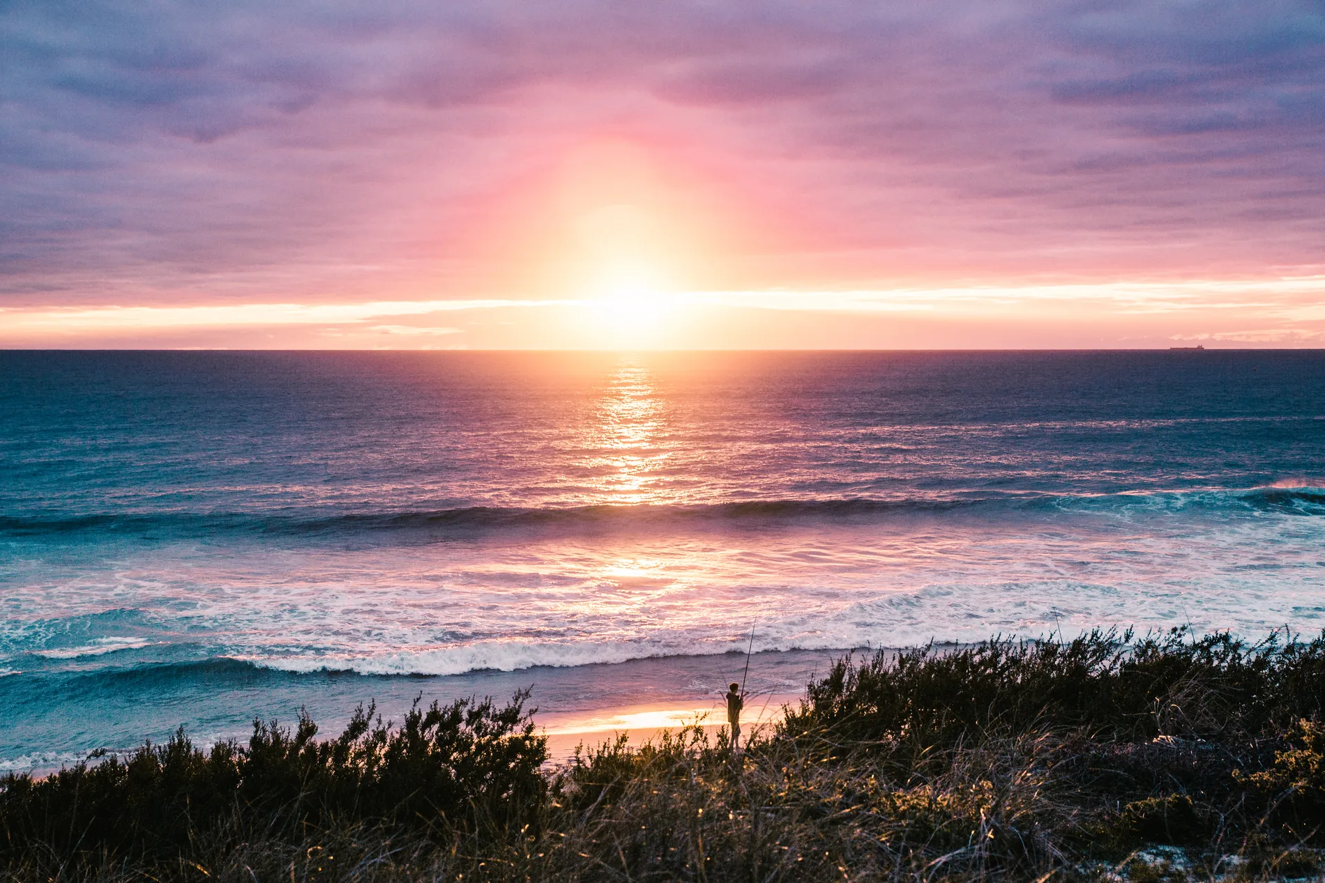 The sun setting into the ocean beyond a low coastal dune and breaking surf