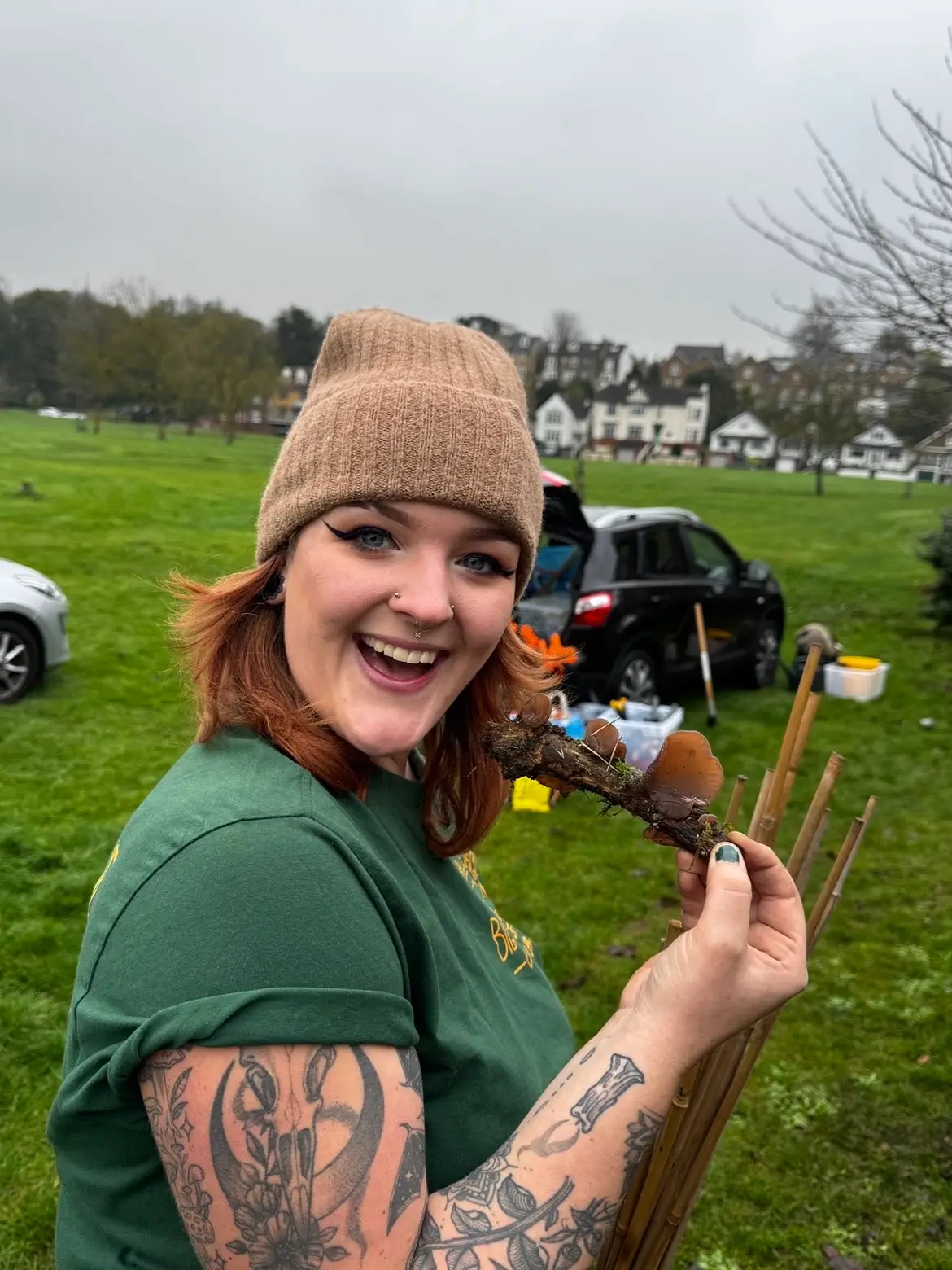 Smiling person with red hair and a beanie holds a stick in a grassy field, with trees and cars in the background.