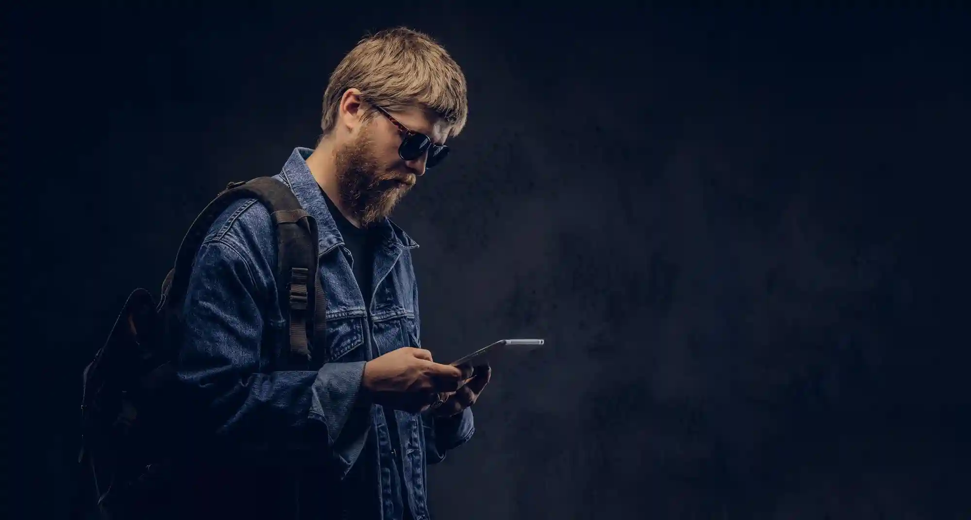 A man wearing a backpack and sunglasses using his smartphone to manage digital tasks while on the go.
