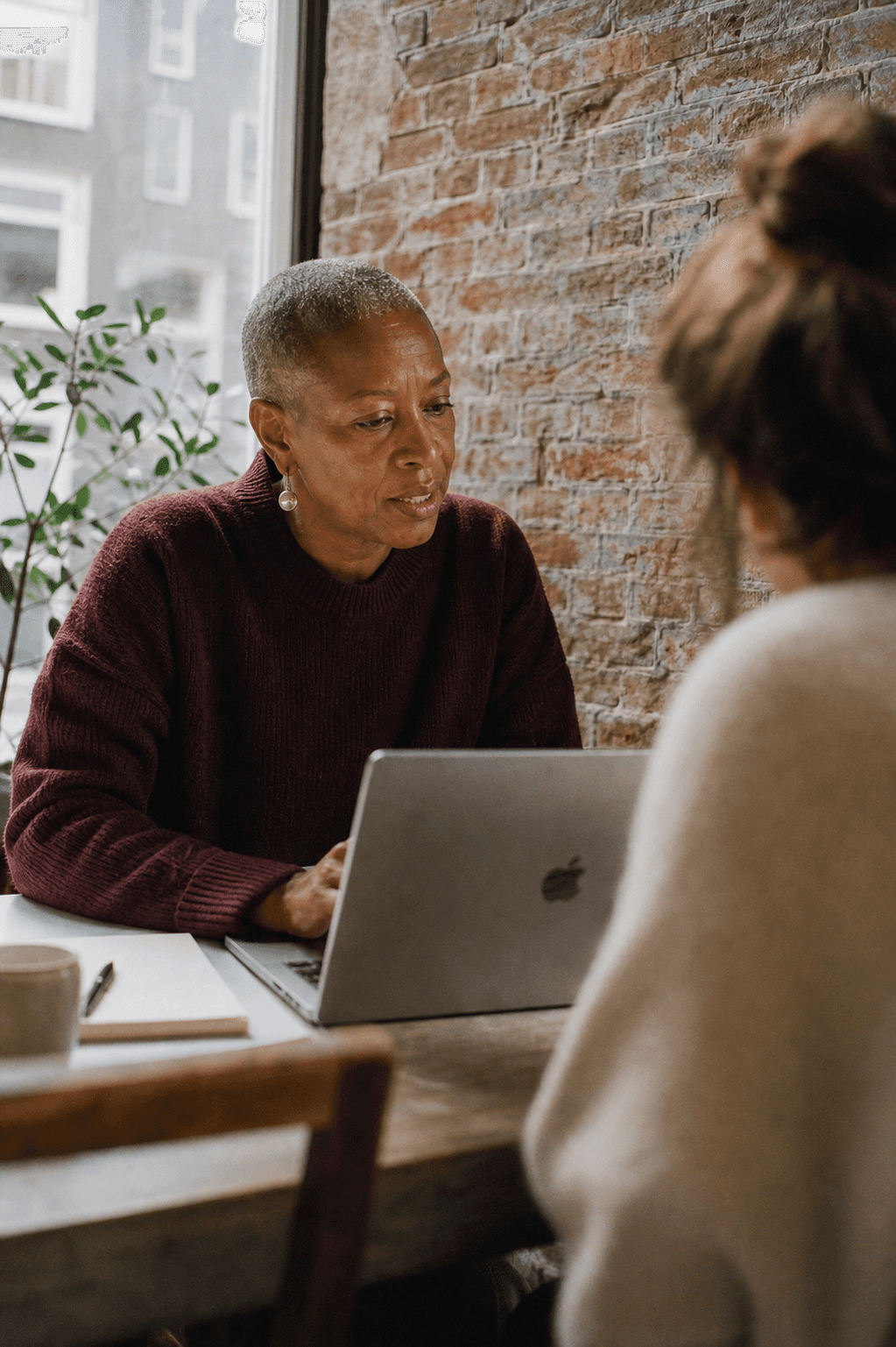 Seated at a brick-walled studio table, reviewing earlier conversations on one open Mac. A colleague is softly out of focus.