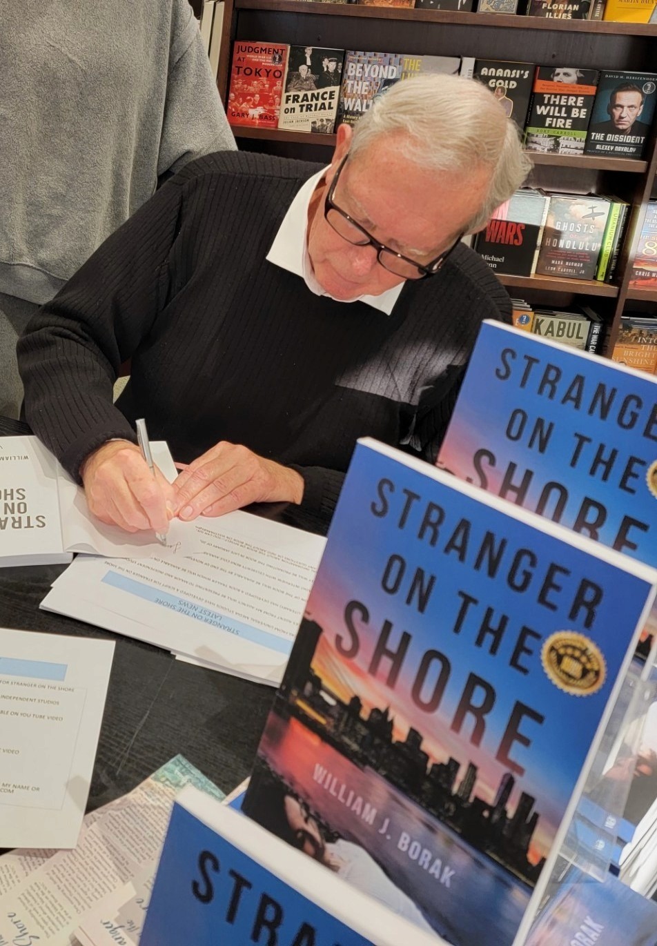 A man with gray hair signs books titled "Stranger on the Shore" at a book signing event. Shelves in the background display various books.