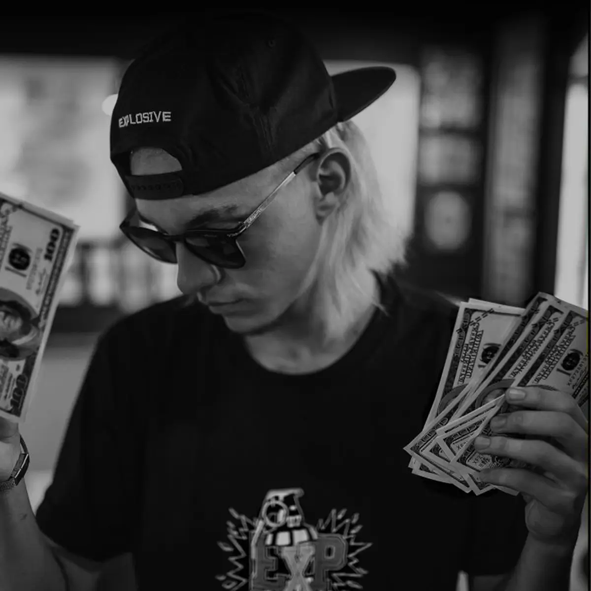 Person wearing a backwards cap and sunglasses, holding several US dollar bills in both hands and looking down at them, shot in black and white indoors.