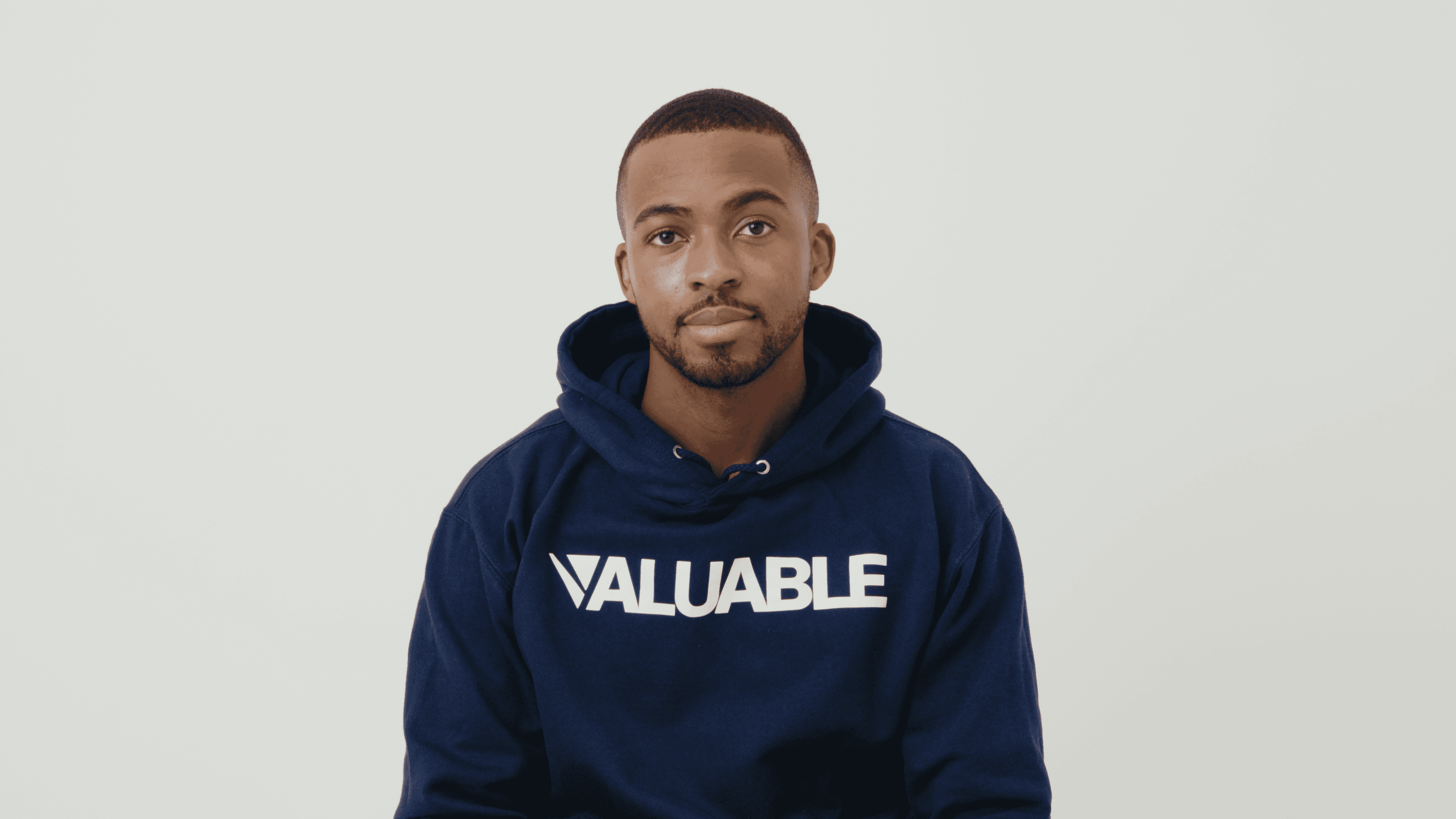 Man in a navy blue valuable hoodie with the word VALUABLE printed on it smiles.