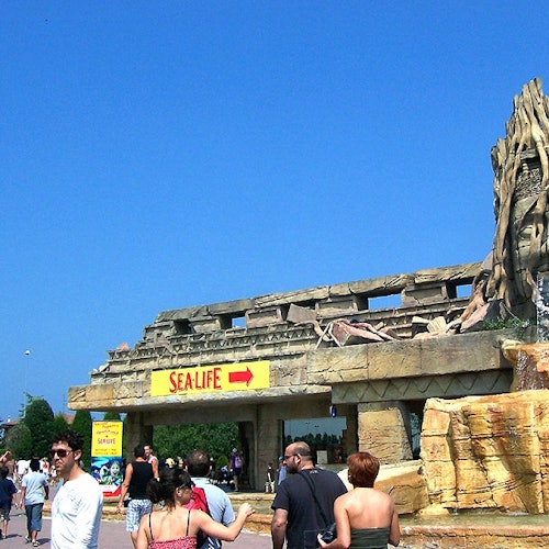 Menschen gehen in der Nähe eines Eingangs vorbei, der mit "SEA LIFE" gekennzeichnet ist, vor einer großen, felsigen Struktur und einem klaren blauen Himmel im Hintergrund.