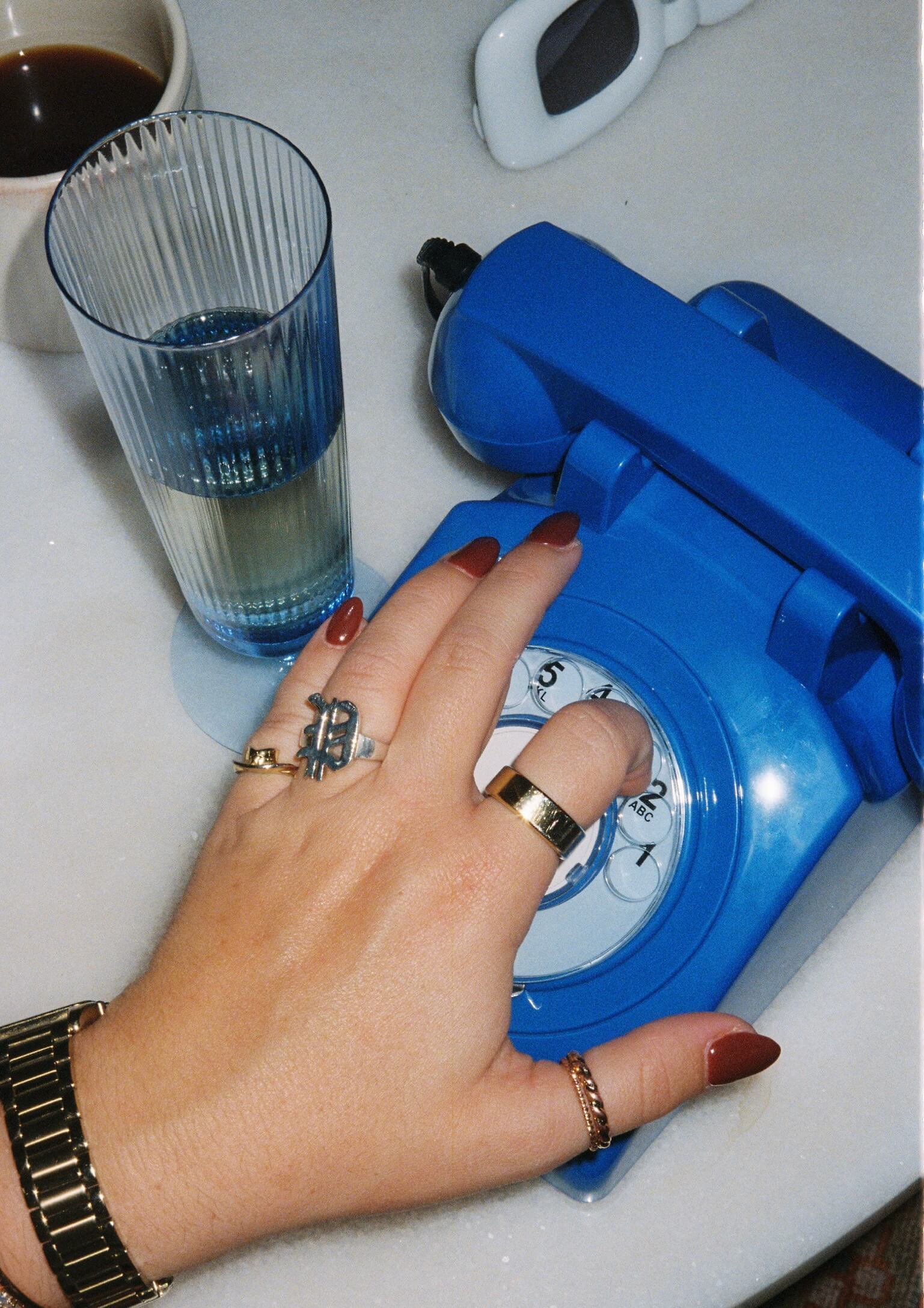 a blue rotary phone sitting on a desk with a glass of water and white sunglasses. A hand is dialing the phone. 