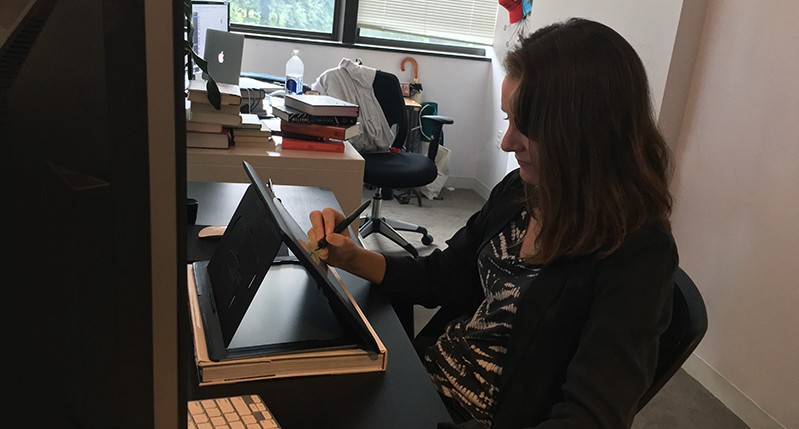A photo of a woman siting in a room and sketching something on a tablet.