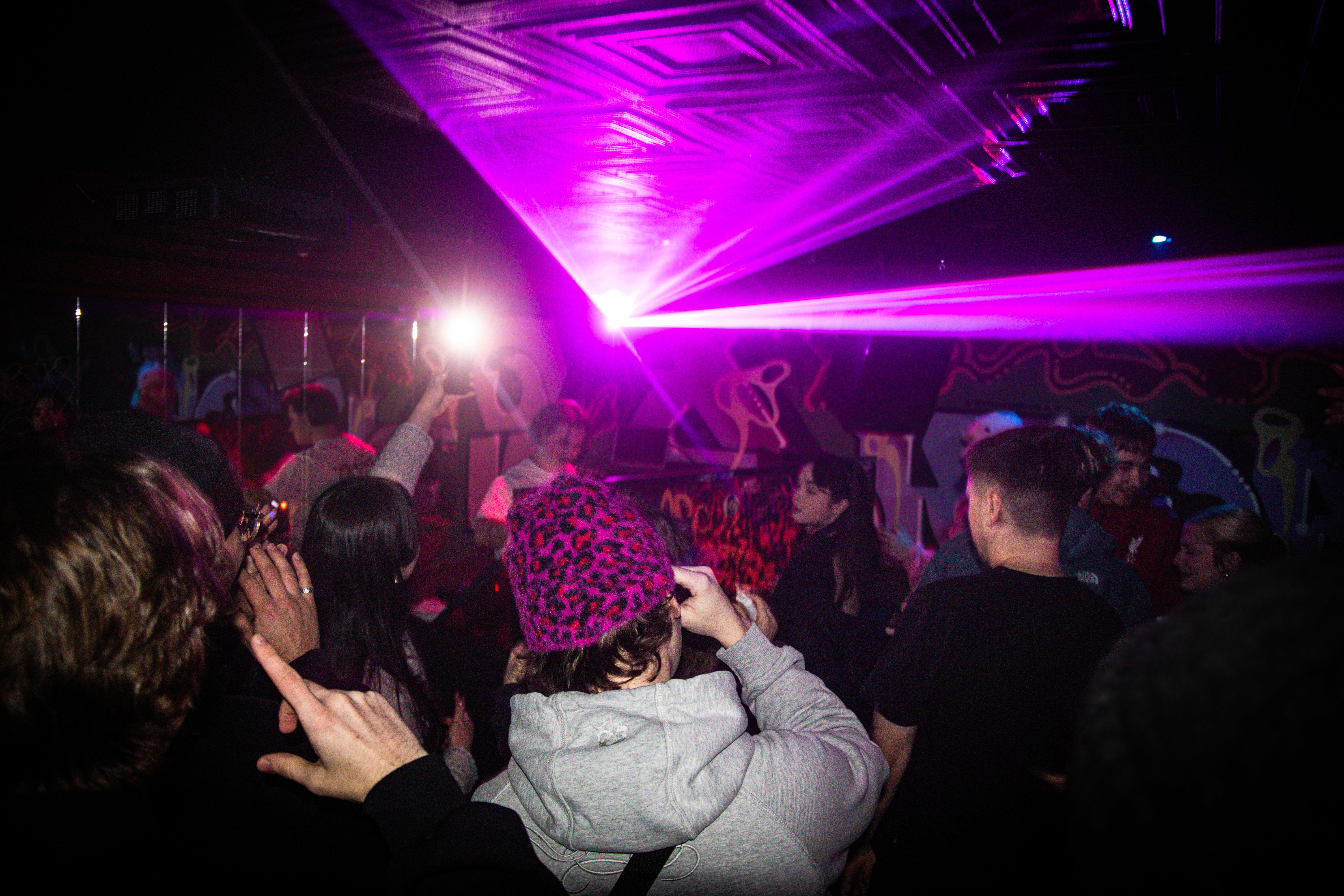 Crowd dancing at a club with hands raised, illuminated by bright pink laser beams cutting through the dark room.