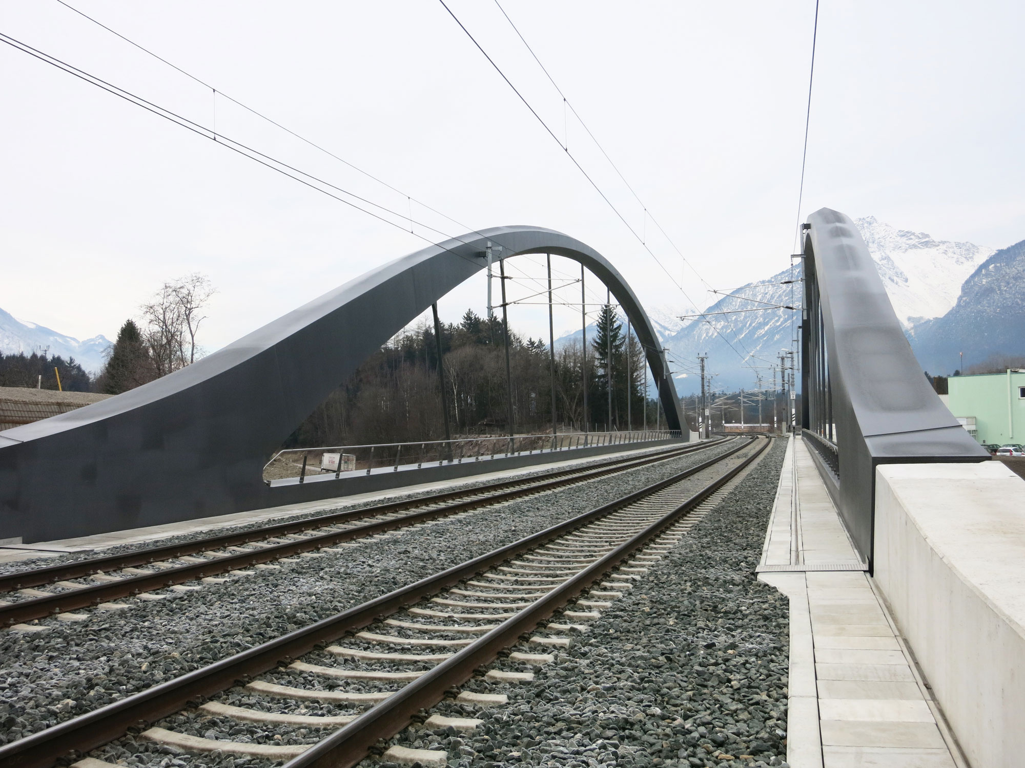 Stahlbogenbrücke mit zwei Eisenbahngleisen, Blick entlang der Bahngleise, Berge im Hintergrund.