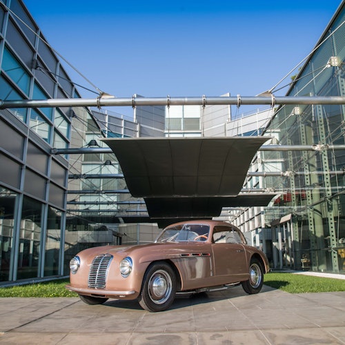 در یک روز آفتابی، یک خودروی قدیمی قهوه‌ای در جلوی یک ساختمان شیشه‌ای مدرن قرار دارد، زیر یک سایبان بزرگ مشکی پارک شده است.