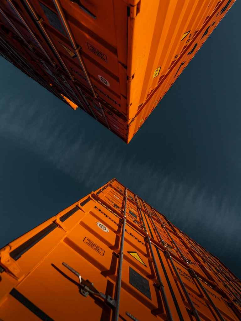 Two towering bright orange shipping containers viewed from below, against a dark sky, representing global logistics and transportation.