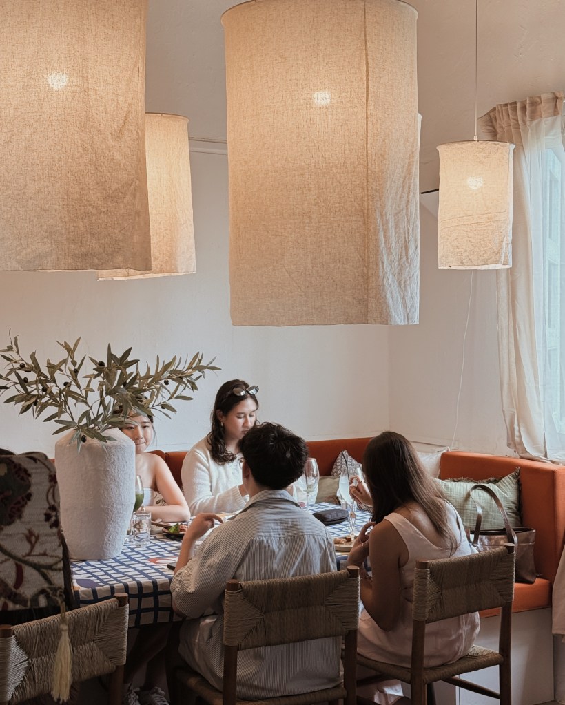 A cozy dining space featuring warm lighting and a relaxed atmosphere, with four people sitting at a table enjoying food and drinks, surrounded by soft furnishings and decorative plants.