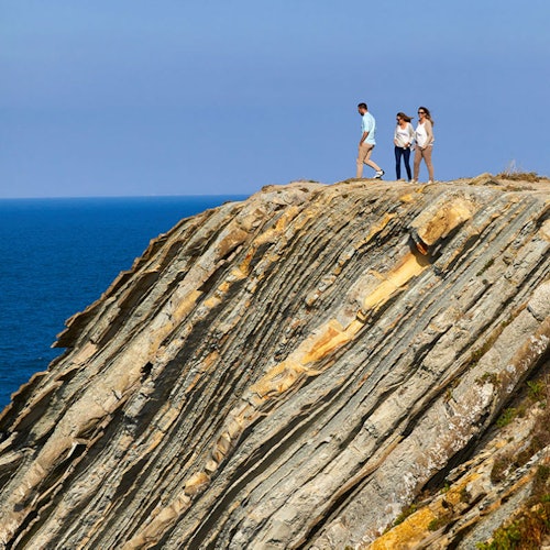 Three people are walking along a rugged cliff edge overlooking the ocean under a clear blue sky.