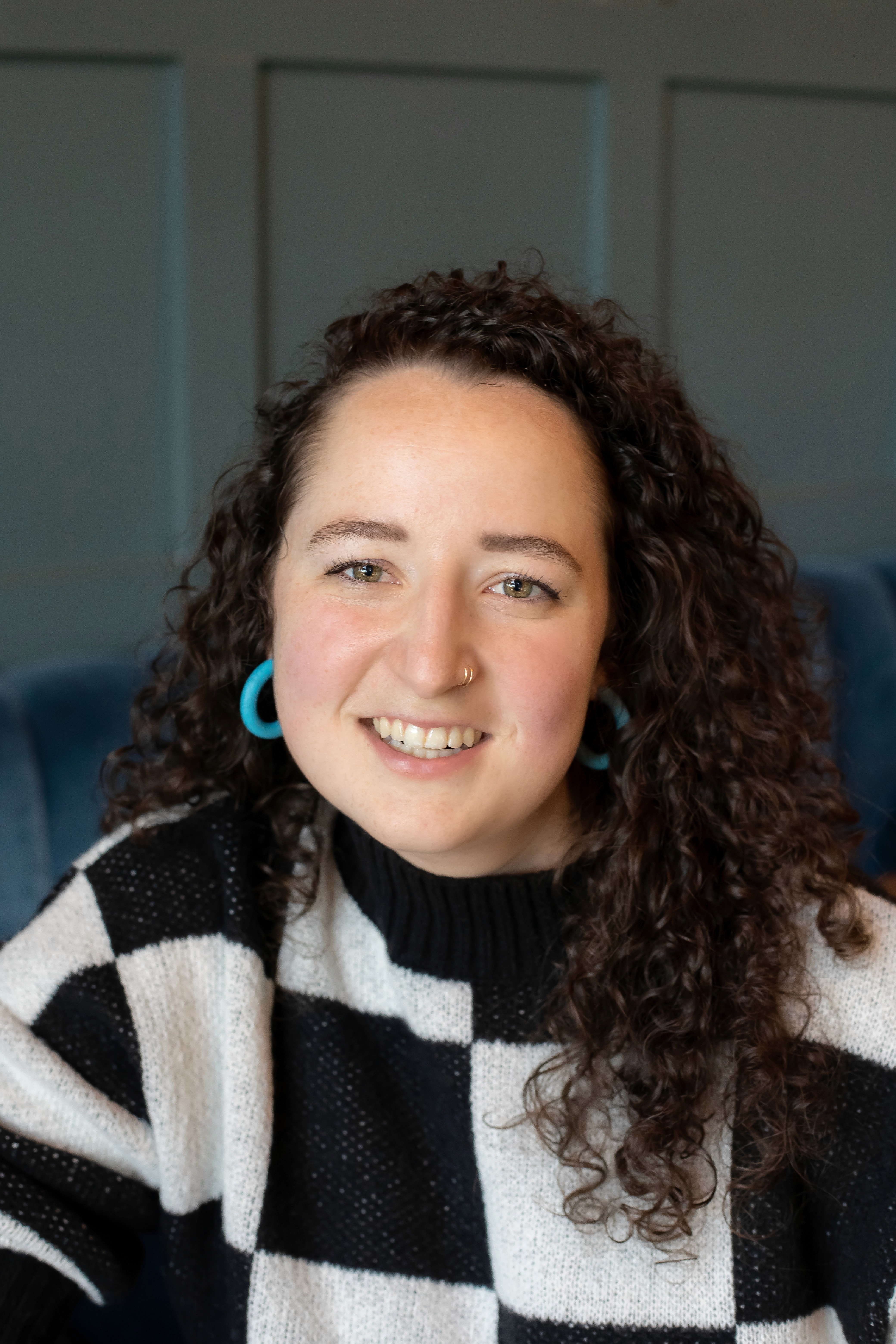 Picture of a woman smiling at the camera. She has dark curly hair, is wearing a black and white checkered sweater, and has electric blue hoop earrings on.