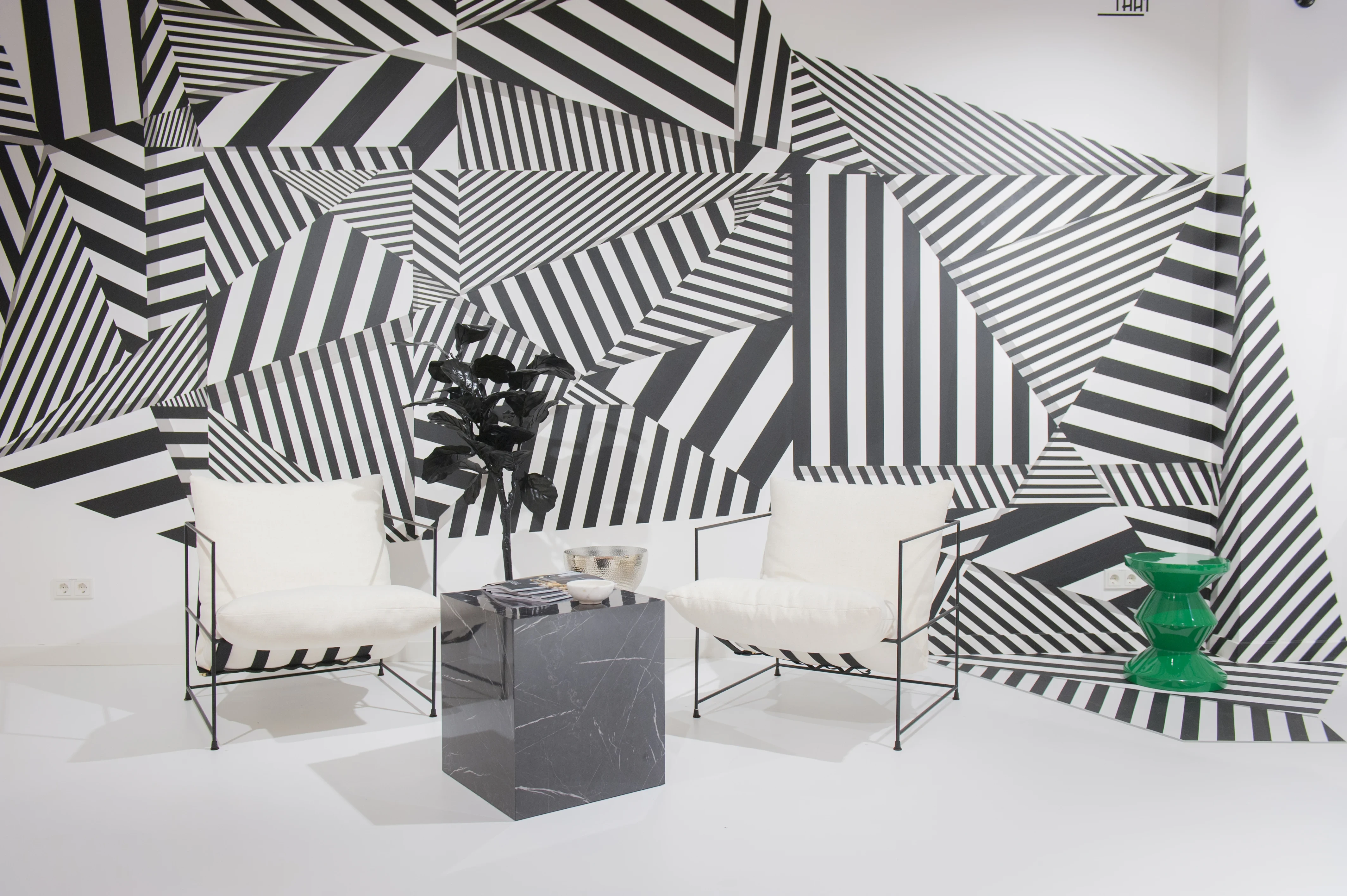 Lounge corner at FREIRAUM SHOP with two white upholstered wire-frame chairs, a black marble block table, a black plant and a bold black-and-white dazzle-pattern wall.