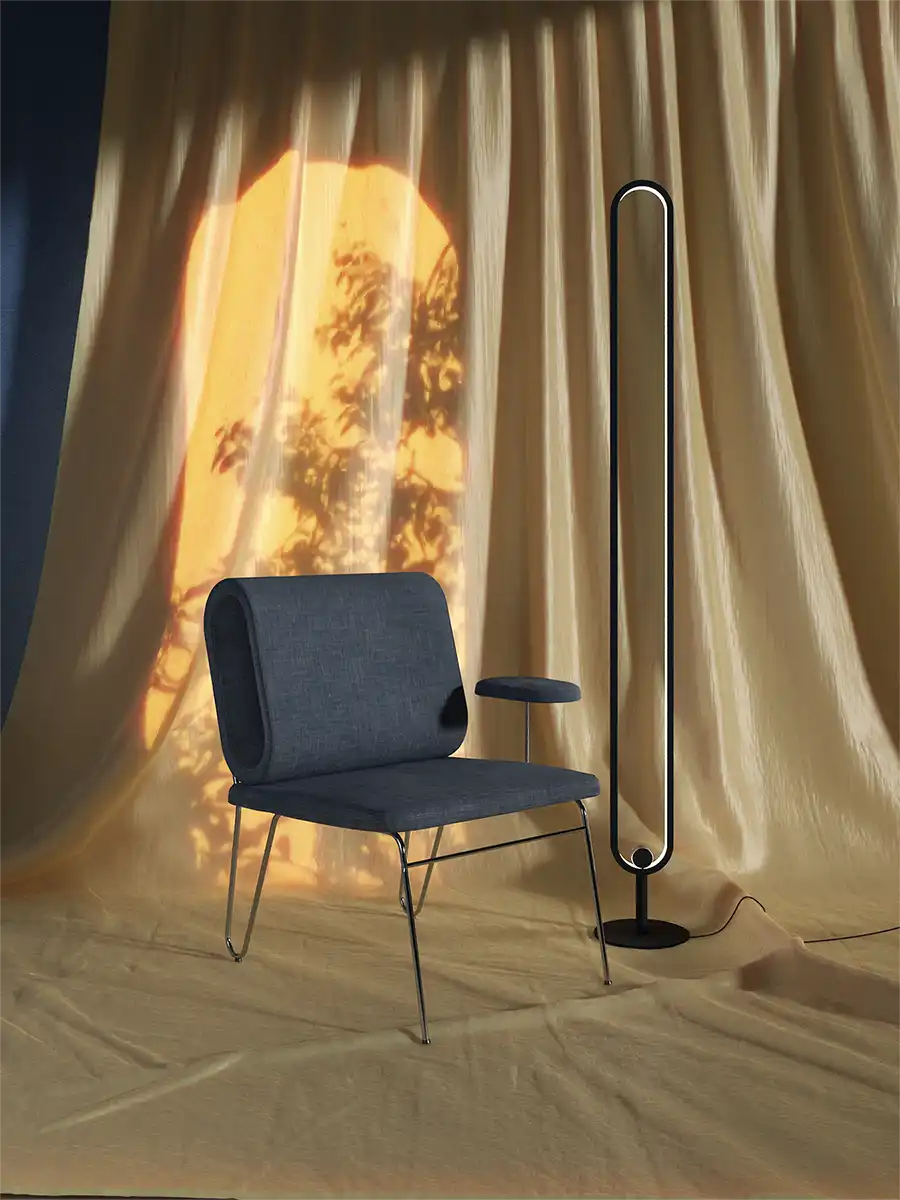 Modern blue upholstered chair with integrated side table next to a tall minimalist floor lamp. Stylish product photography with warm sunlight and curtain shadows.