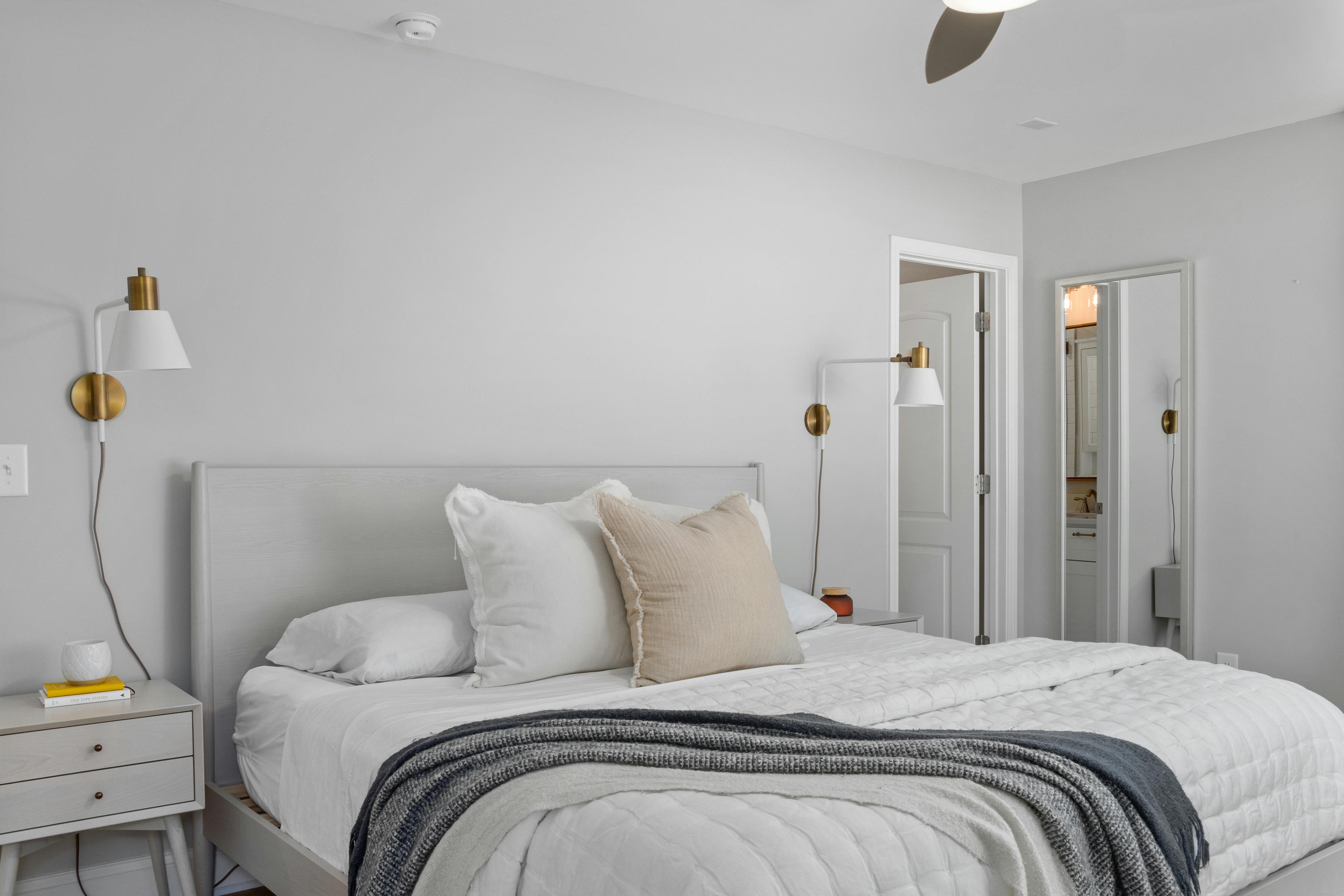 A serene bedroom with a white bed frame, soft linens, and neutral-toned cushions. Elegant wall-mounted lamps add a touch of sophistication, and a doorway leads to an en-suite bathroom