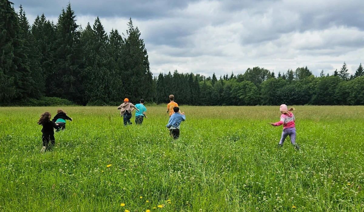 Children run freely across a wide green meadow at Rooted Northwest, with trees lining the horizon.