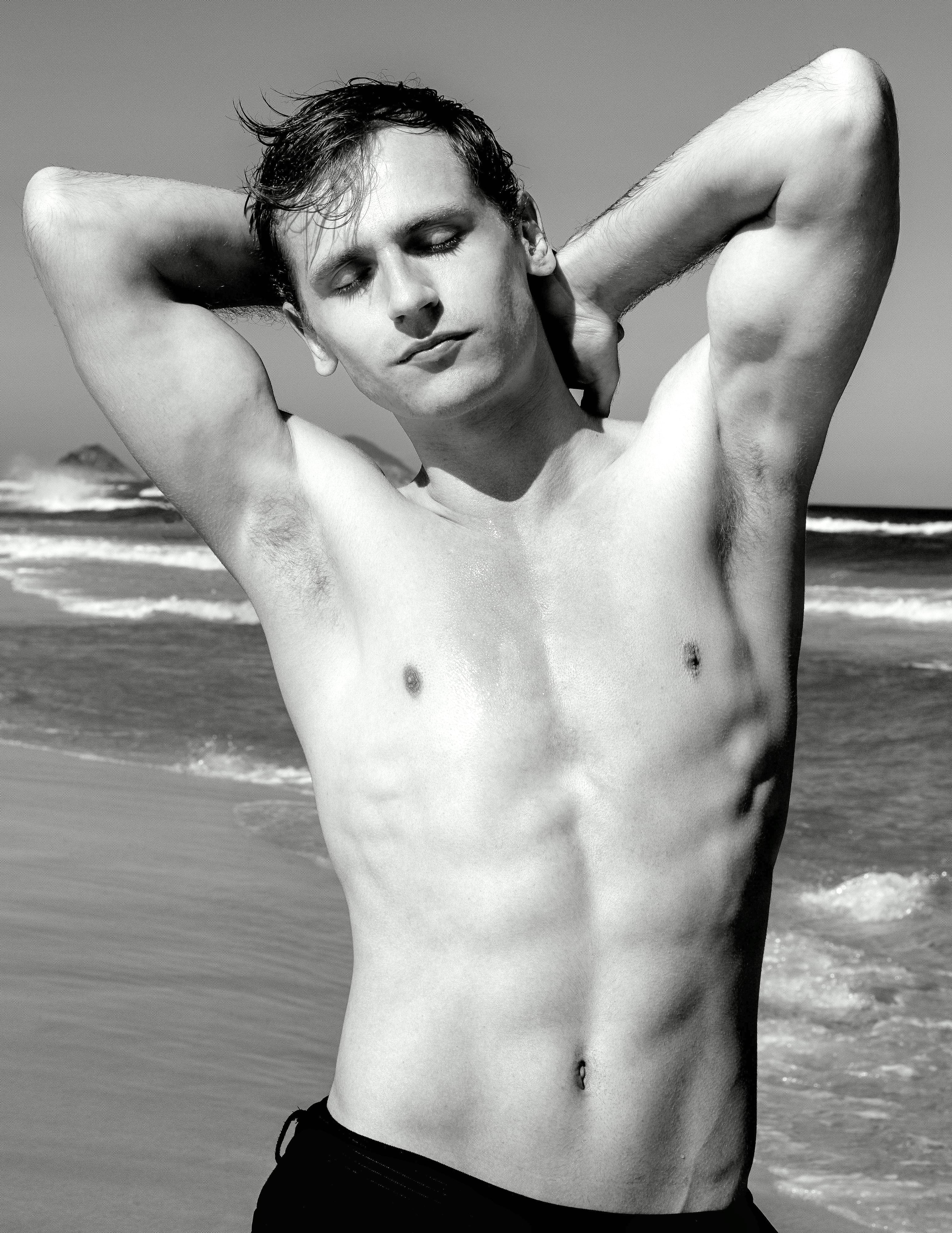 Black and white image of a shirtless man with closed eyes and hands behind his head, standing on a beach with the ocean waves in the background