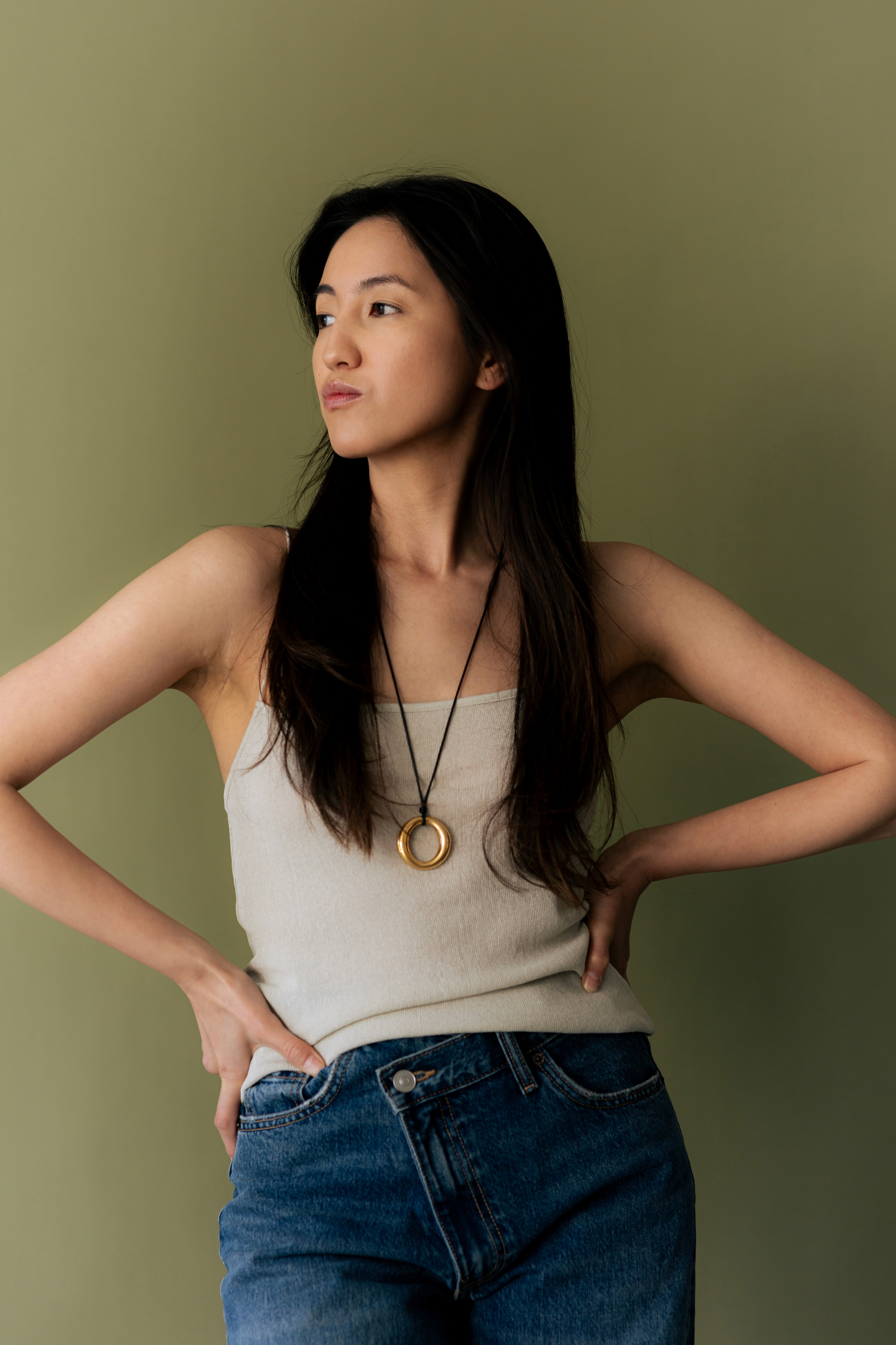 Woman with long dark hair, hands on hips against an olive green background.