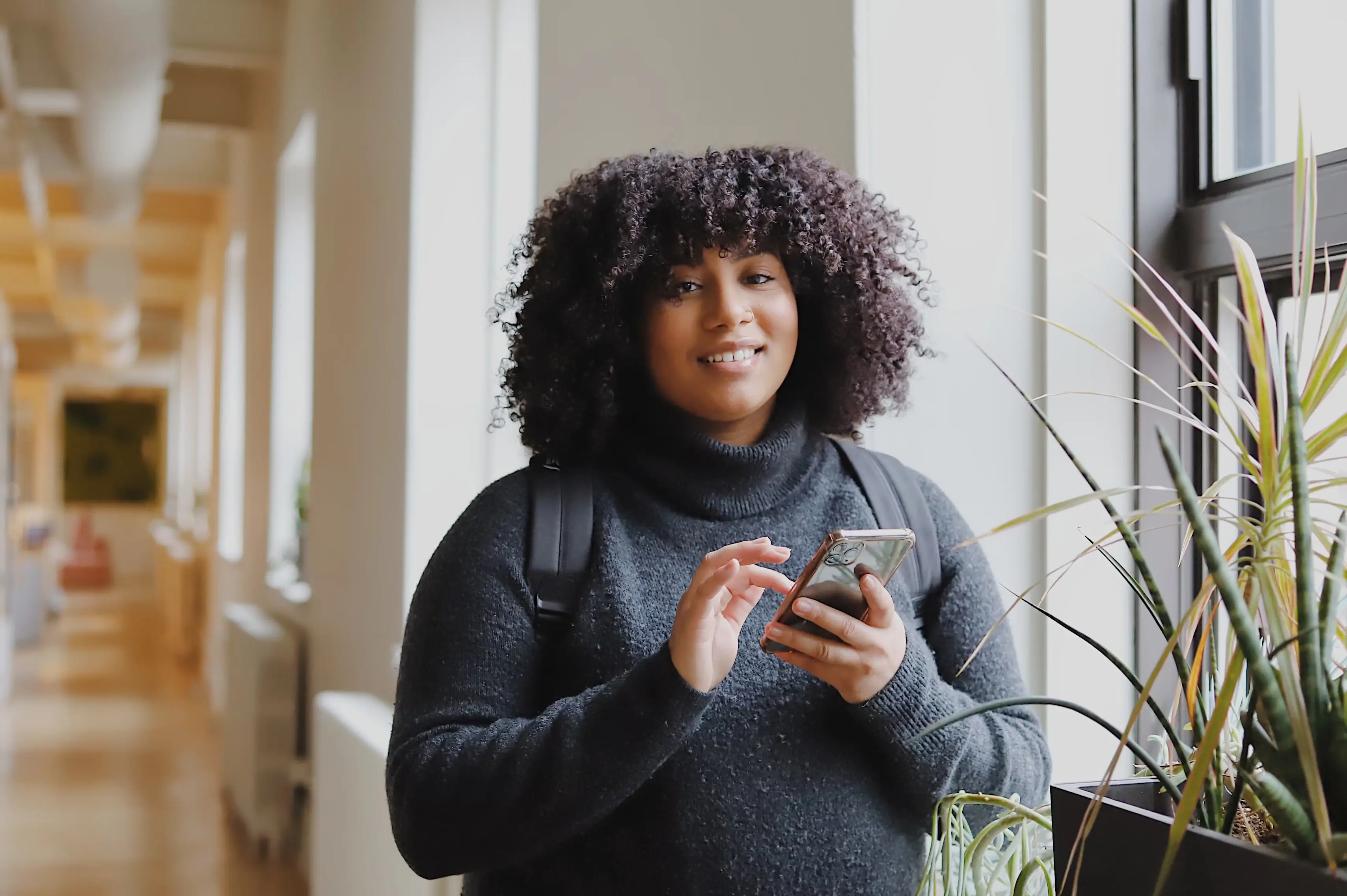 Lady with curly hair, wearing a backpack smiling holding her phone in an office space.