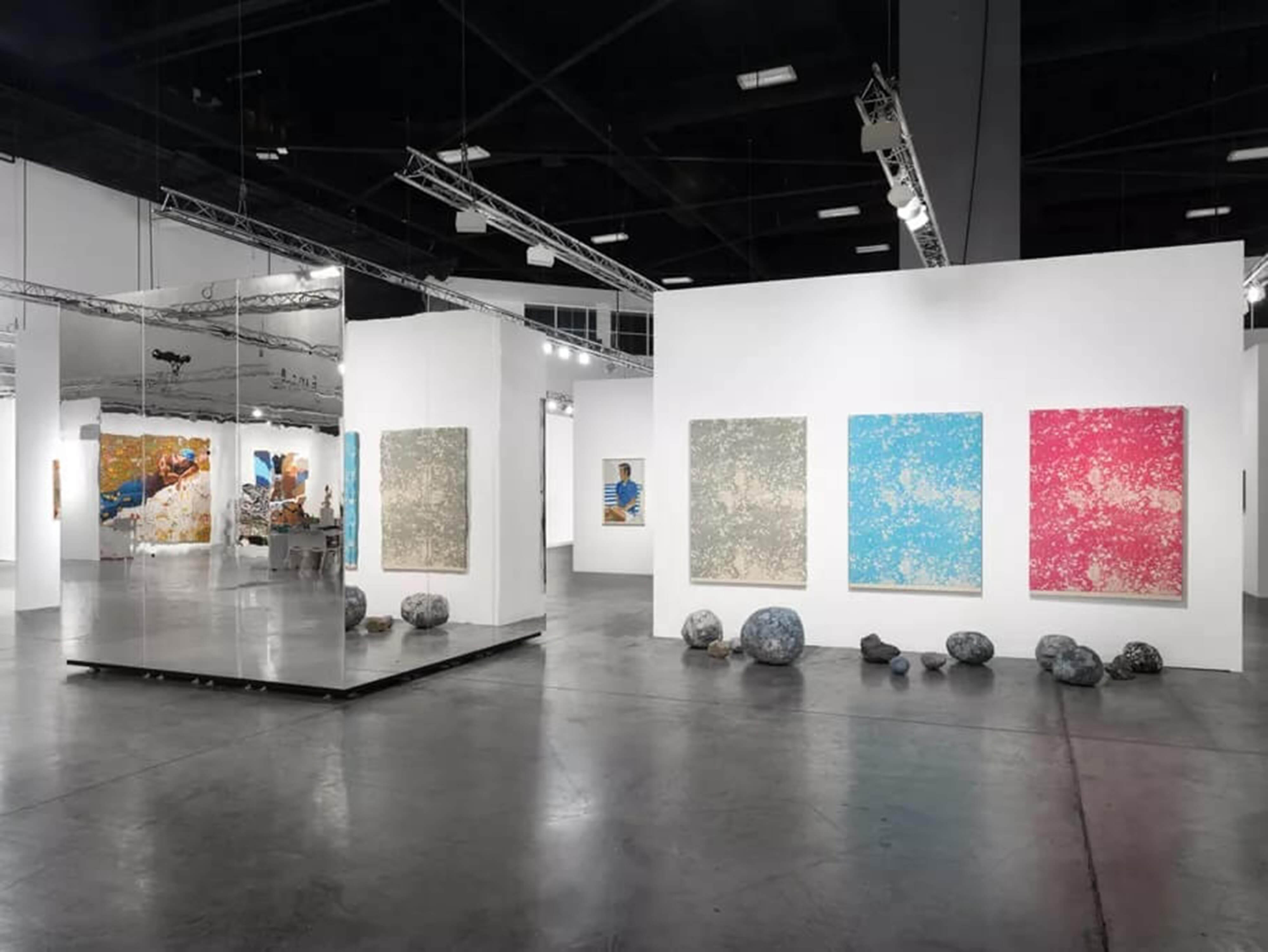 Art fair booth with a mirrored installation, colorful paintings, and stone-like sculptures placed on the floor.