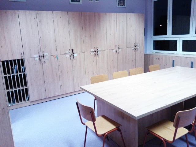 Funkcjonalne szafki skrytkowe i regały z jasnego drewna wykonane dla biura lub placówki edukacyjnej, wraz z dopasowanym stołem konferencyjnym.