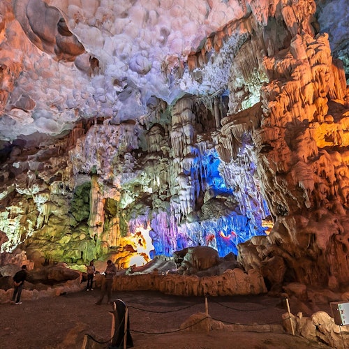 En lyst oplyst grotteinteriør med farverige klippeformationer og stalaktitter. Flere mennesker står og beundrer landskabet.