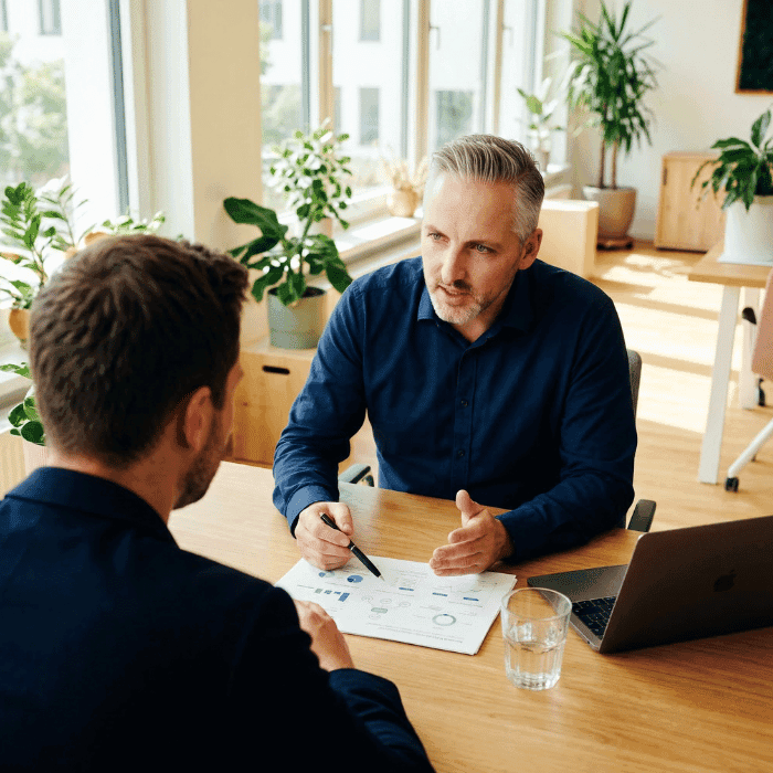 Versicherungsmakler Hannes Weindorf erklärt einem Geschäftsführer die betriebliche Altersvorsorge am Schreibtisch mit Dokumenten und Diagrammen