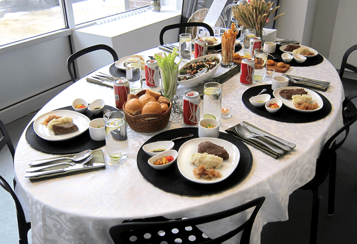 A white tablecloth covers a dining table set with black placemats and silverware. Four place settings are visible, each with a white plate, silverware, and glass of water. Black chairs surround the table. A centerpiece of bread rolls in a woven basket sits in the middle of the table. Condiments, including ketchup and mustard, are in small bowls. Four cans of soda are placed near the center. A small bowl of sliced tomatoes is visible near the bottom left corner. A vase of tall grass or dried plant material is in the background. A window is visible on the left side of the image.