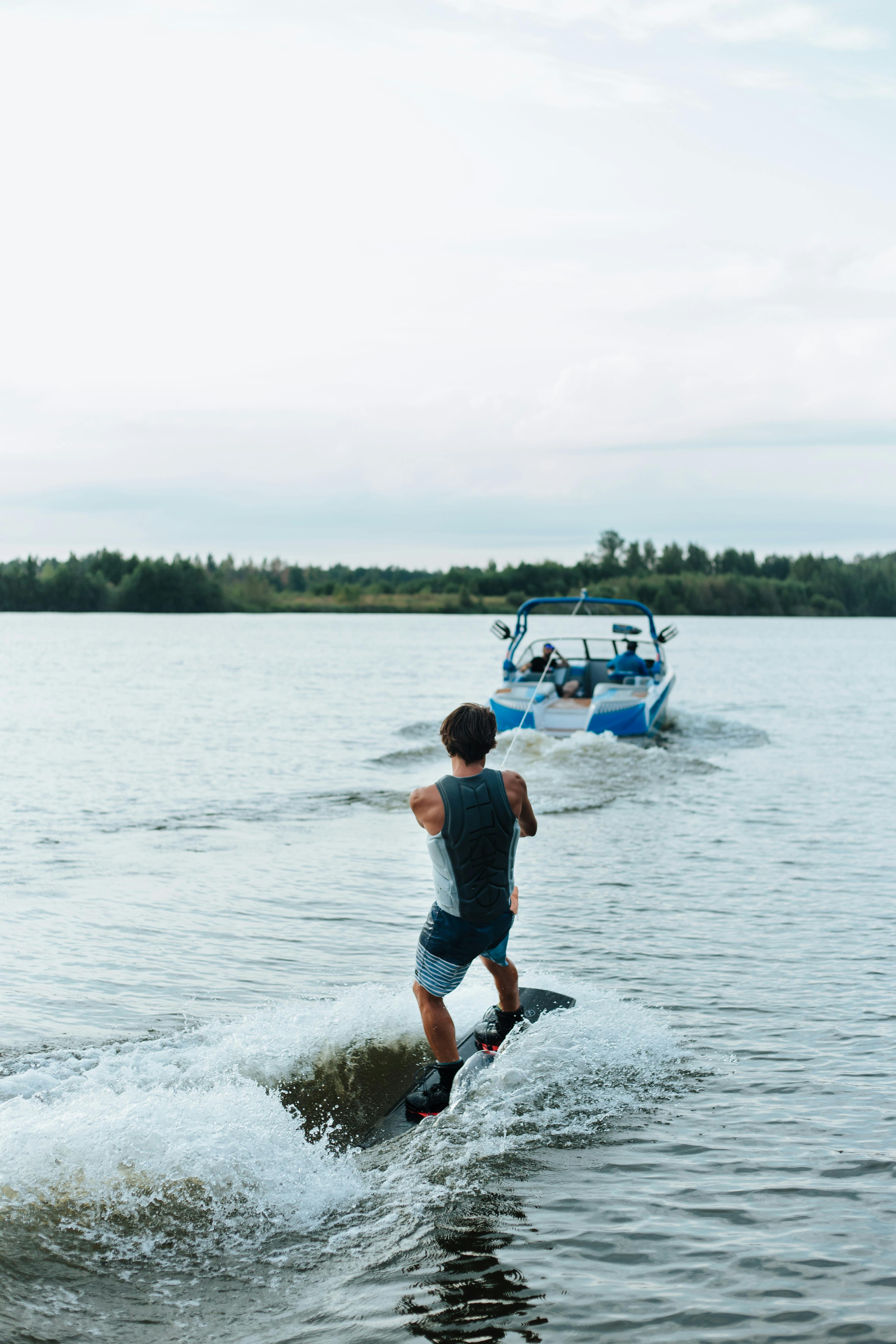 A person in a wetsuit wakeboards behind a blue and white speedboat on a calm lake, surrounded by lush green trees and a cloudy sky.