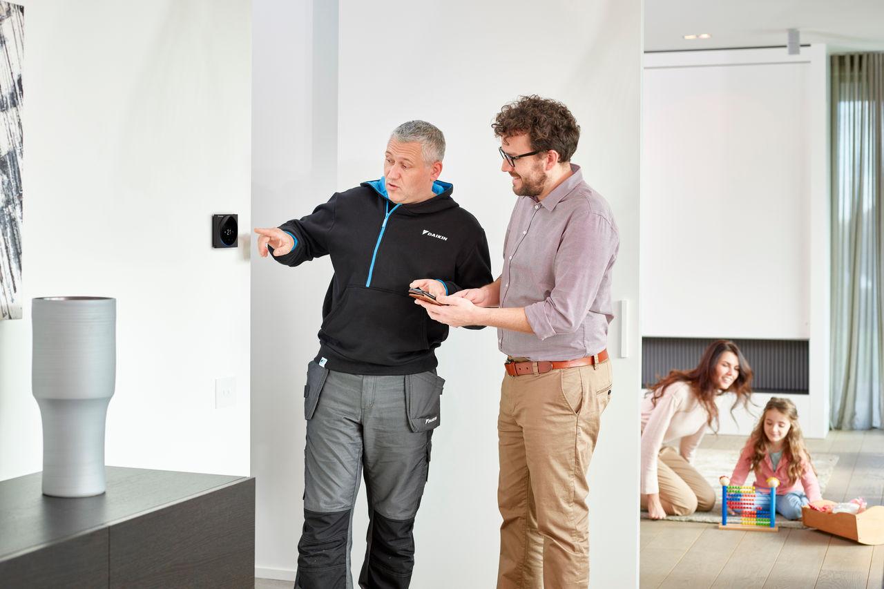 A heat pump engineer explaining controller settings to a homeowner in a modern UK living room while a family relaxes in the background.