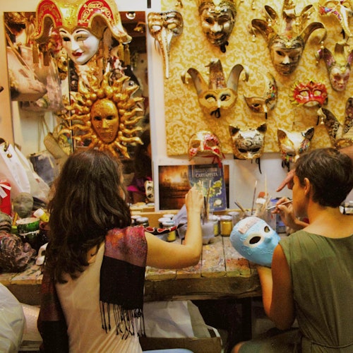 Due persone sono sedute a un tavolo ingombro, mentre dipingono maschere in un laboratorio decorato con varie maschere veneziane sulla parete.