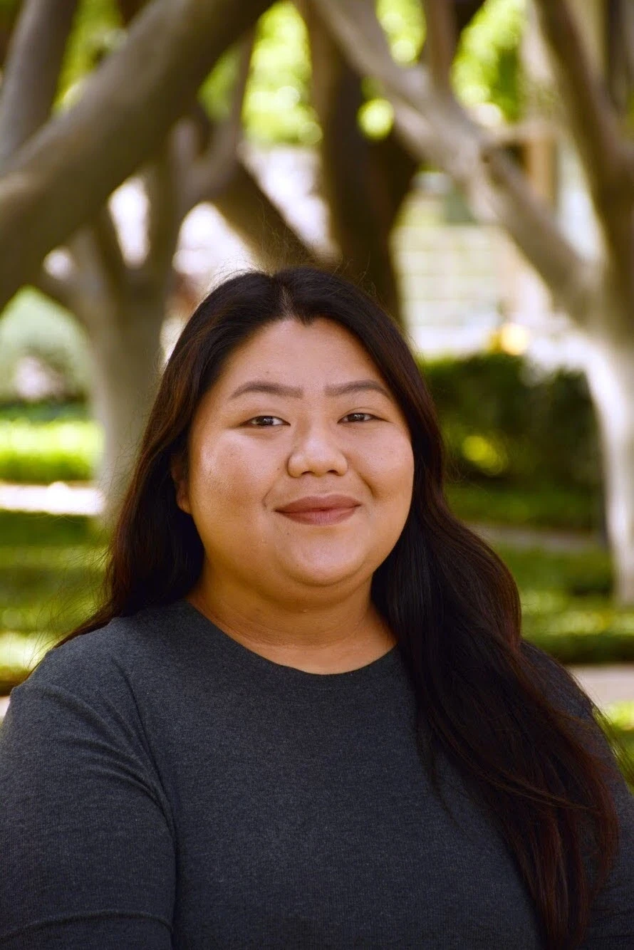Uma pessoa com cabelo longo e escuro e uma blusa cinza, em pé ao ar livre diante de um fundo desfocado de troncos de árvores e folhagem verde brilhante, sugerindo um ambiente natural sereno.