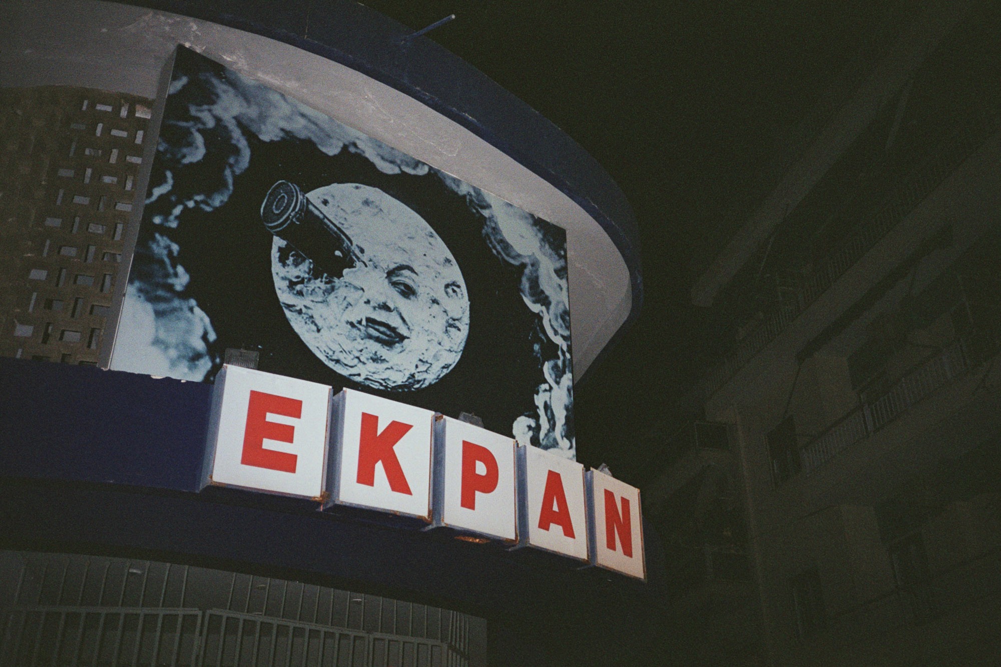 A vintage-style sign on a building facade features a whimsical illustration of the moon with a telescope protruding from it, above the bold red letters spelling "EKPAN."