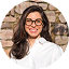 Jeune femme aux longs cheveux bruns, souriant largement avec des lunettes, devant un mur de briques.