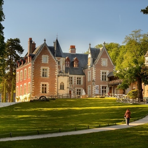 A person walking on a path toward a large, historic, red-brick mansion surrounded by green lawns and trees on a sunny day.