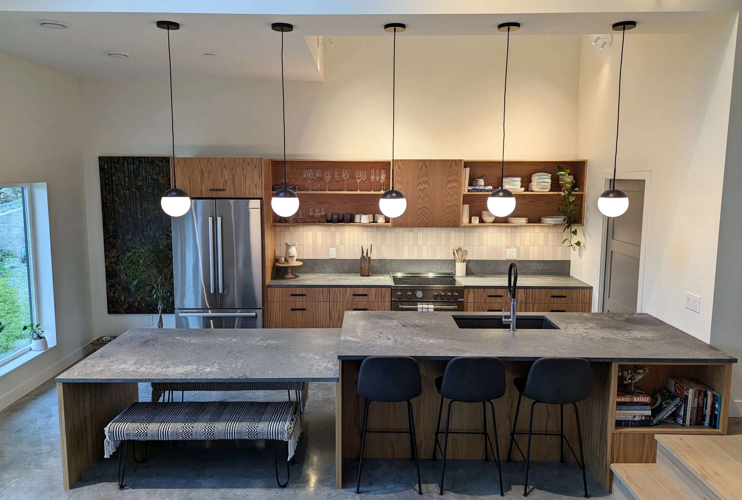 A modern, glass-walled house with warm interior lighting is perched on a rocky cliff overlooking a calm sea at sunset, under a clear blue sky.