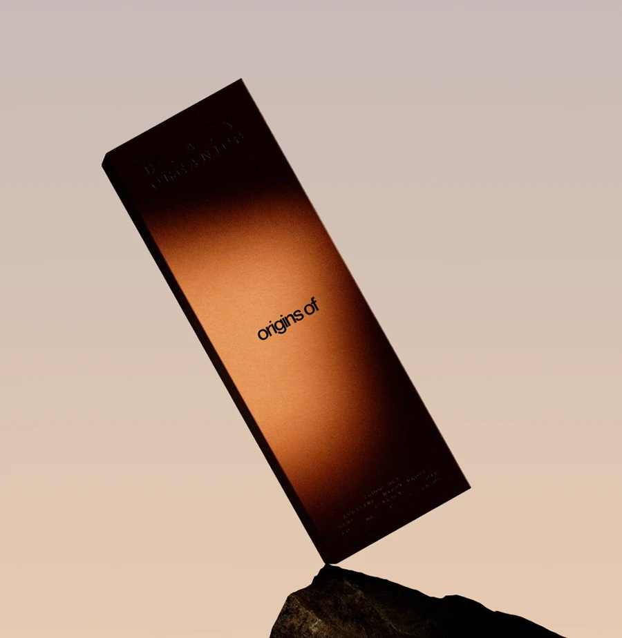 A tall Origins Of box in a warm amber-to-black gradient, balanced at an angle on a dark rock against a soft peach sky.