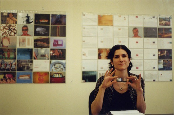 A photo of a woman holding some kid of 3d logo while sitting in front of a wall with pictures and paper articles on it..