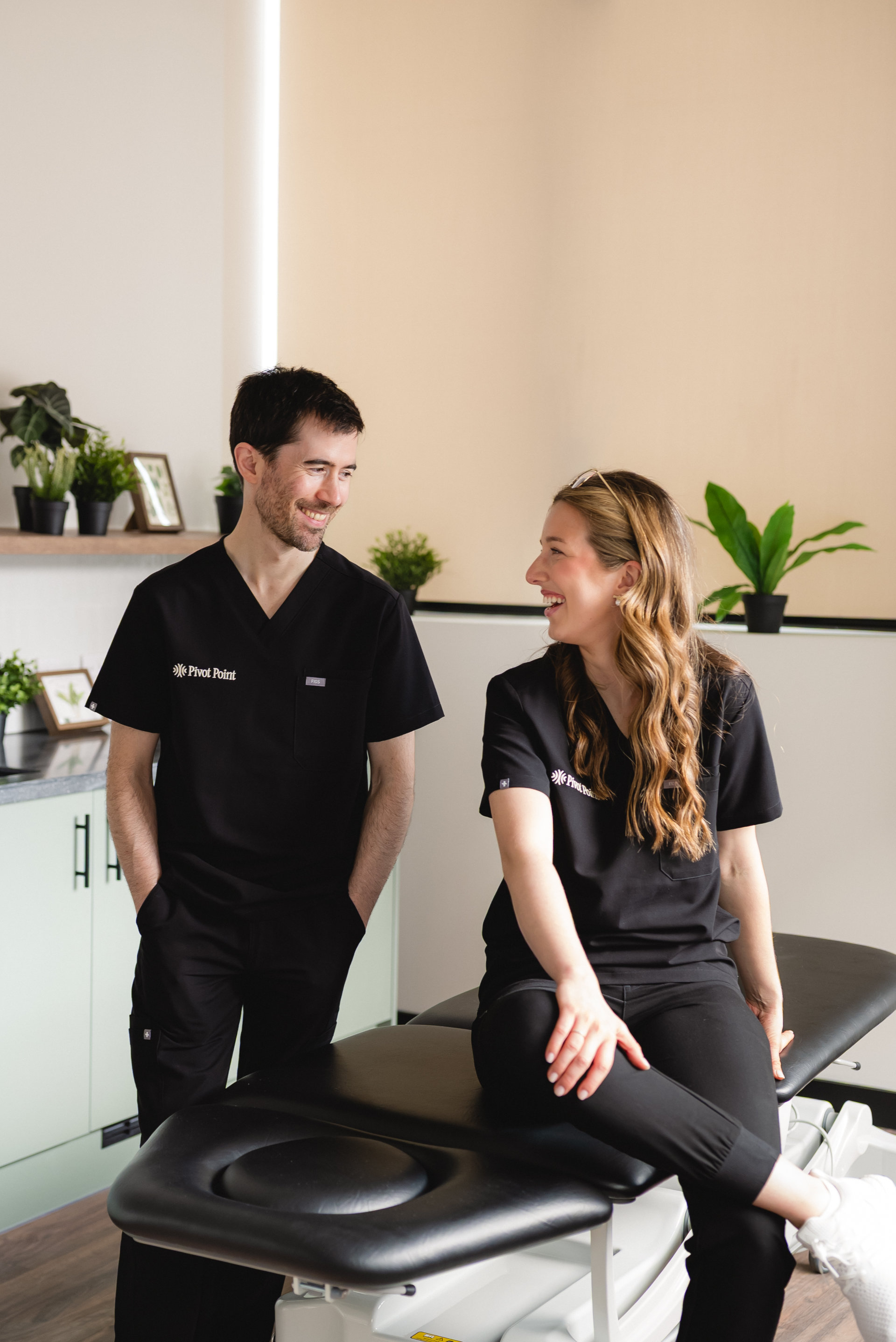 Two physiotherapists in uniform in a treatment room, facing each other and smiling