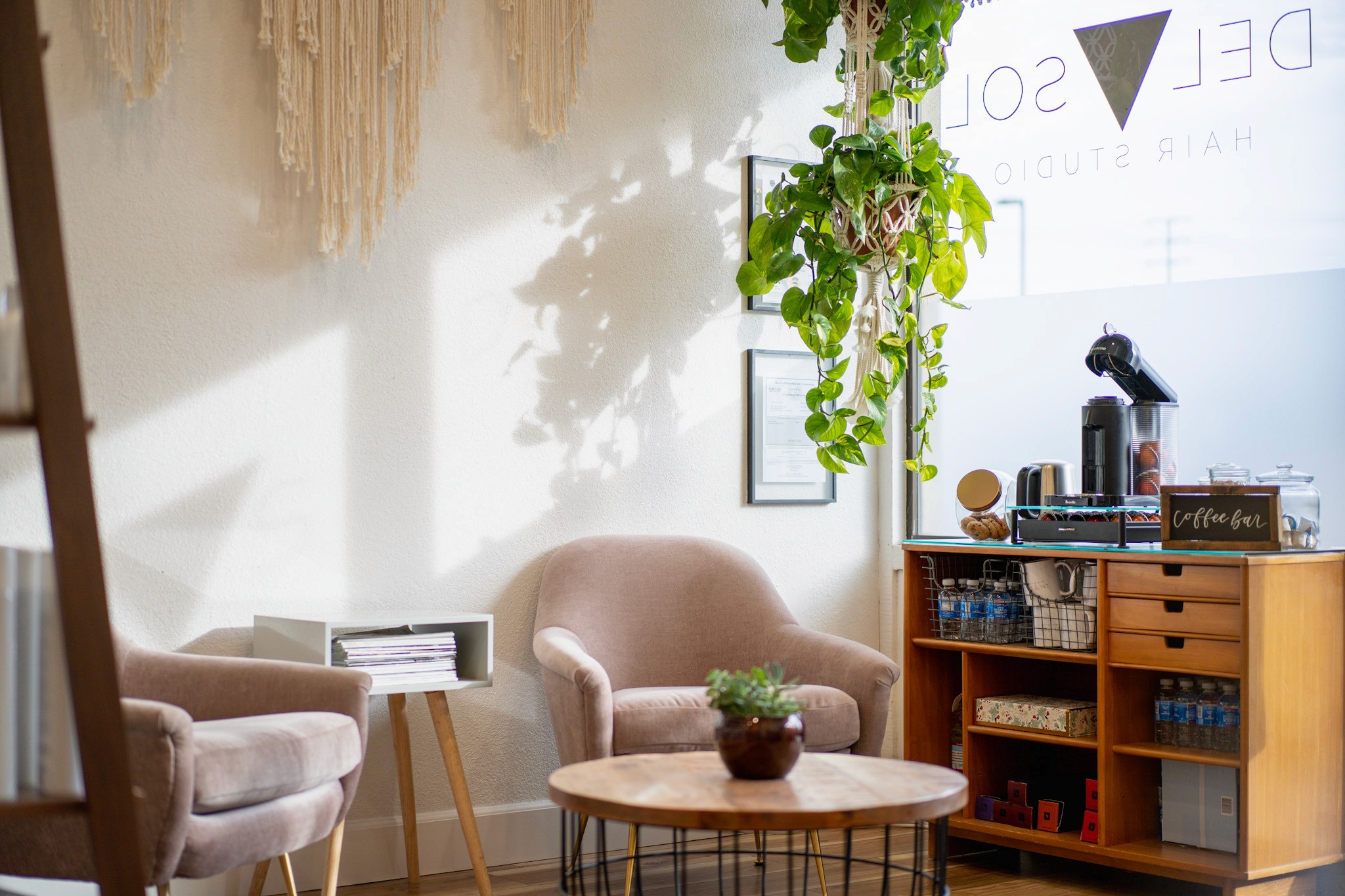 Cozy coffee bar at the entrance of Del Sol Hair Studio, featuring soft seating, hanging greenery, natural light, and a curated beverage station for guests.