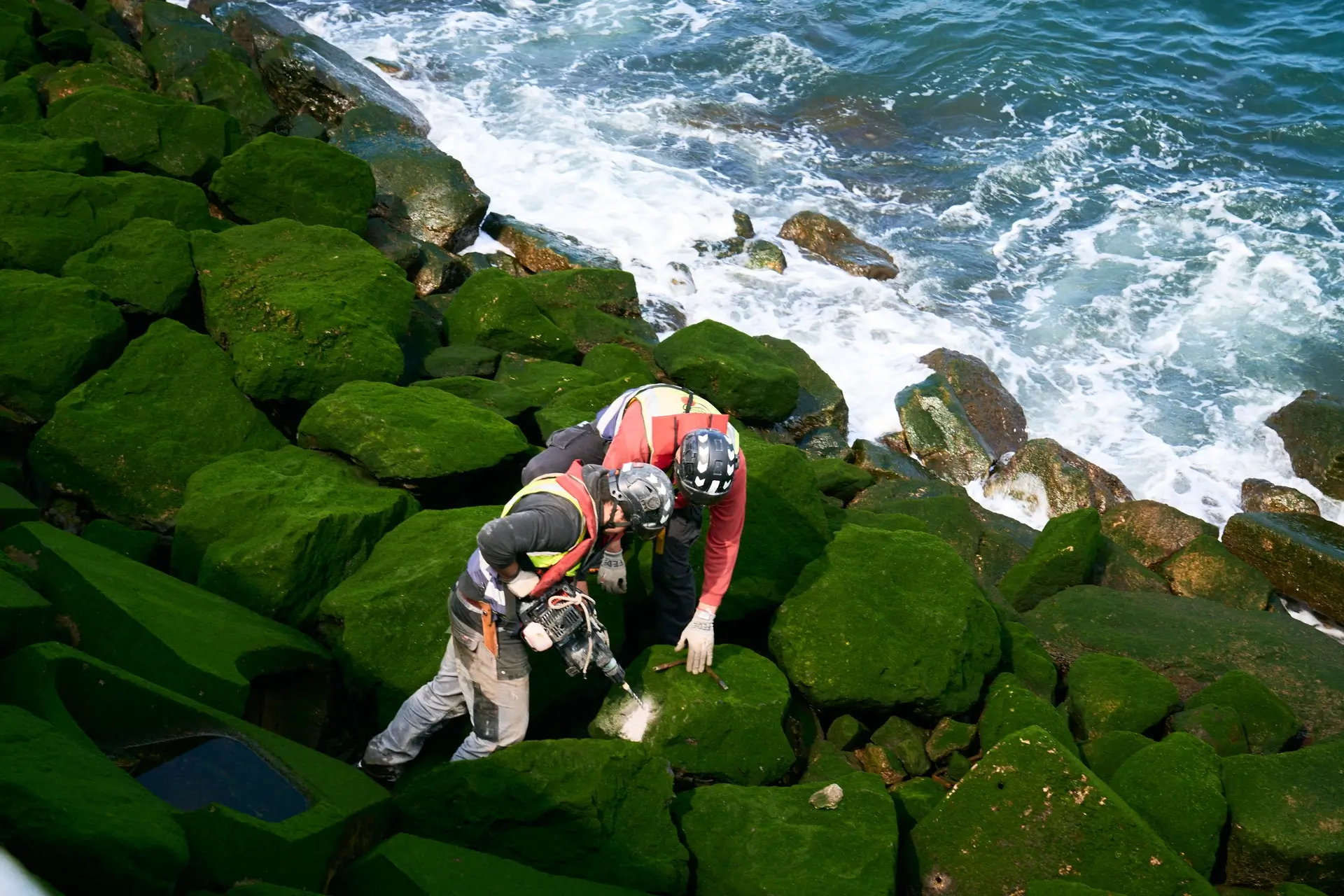 Two workers install monitoring equipment on algae-covered coastal armor blocks in the tidal zone beside the ocean.
