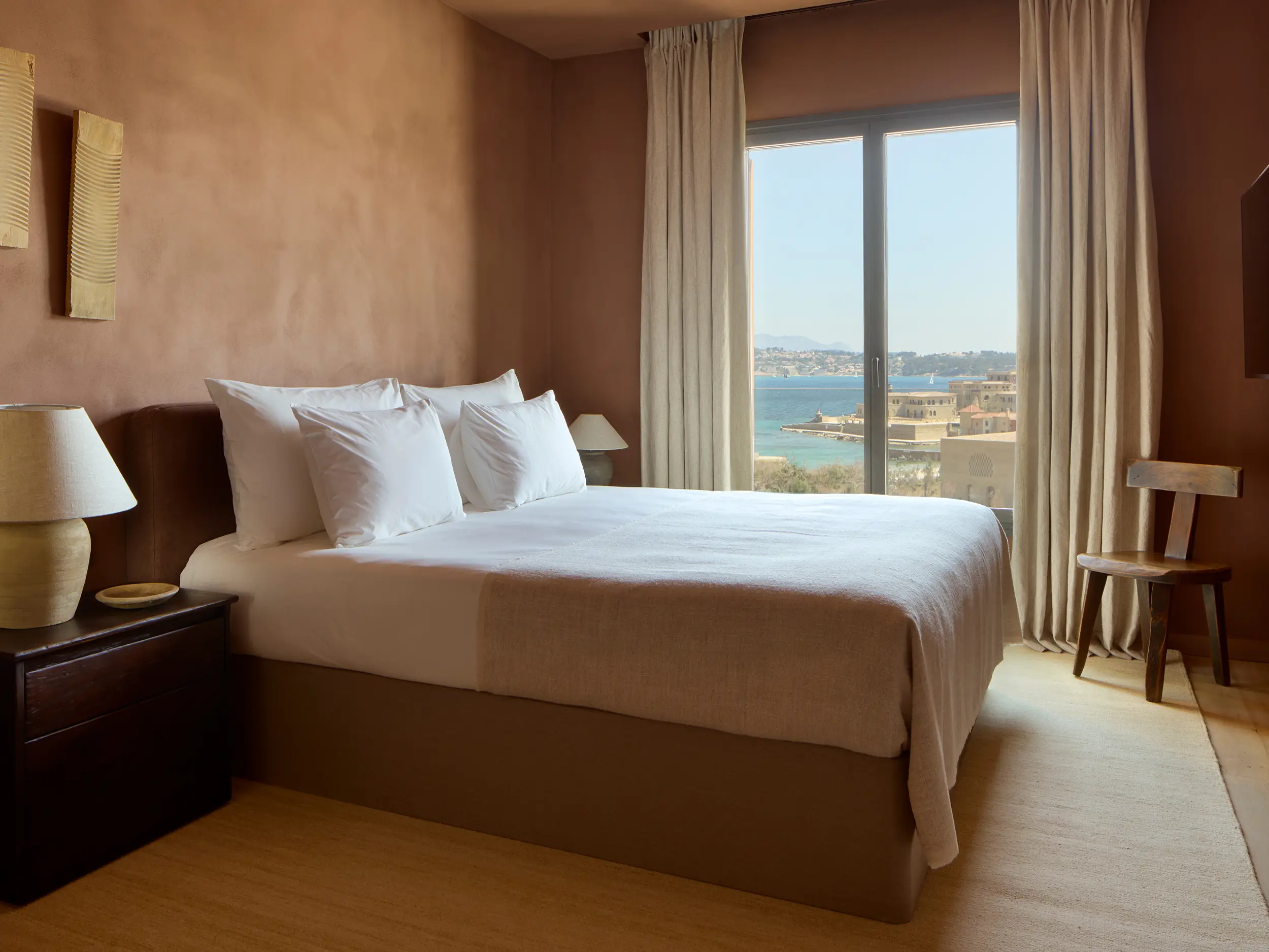 Helles, minimalistisches Hotelzimmer mit Blick auf das Meer durch ein großes Fenster. Ein gemütliches Doppelbett mit weißen Laken steht vor einer terracottafarbenen Wand, daneben ein dunkler Nachttisch und ein rustikaler Holzstuhl.