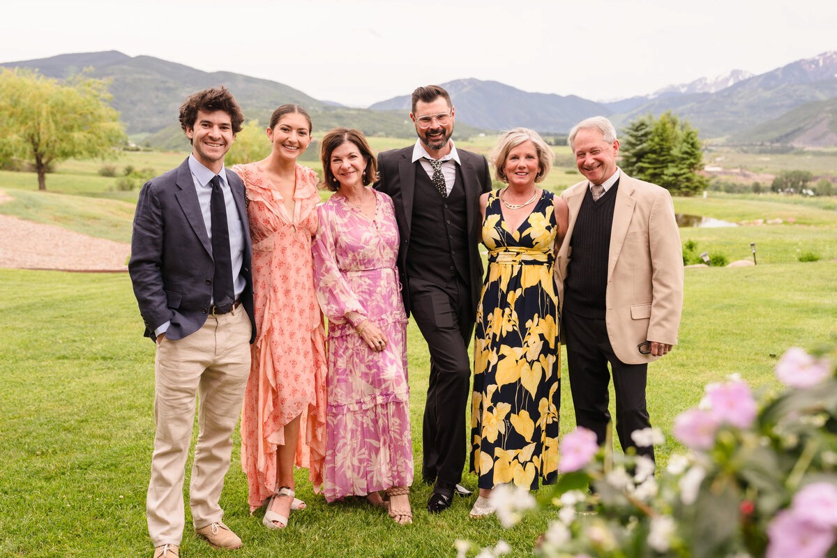 Six guests in colorful dresses and jackets posing on a lawn with mountains behind them