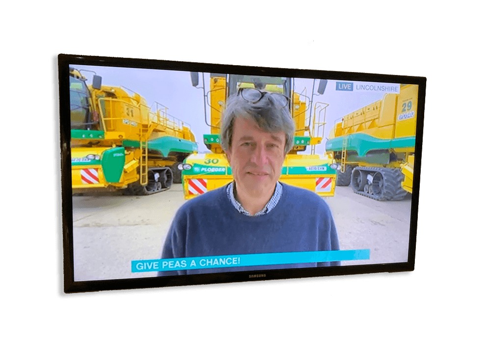 Flat screen TV with ablack surround showing bright yellow farm vehicles in the background. Stephen Francis, wearing a mid blue sweater, is standing in the foreground. Caption reads 'Give peas a chance!'