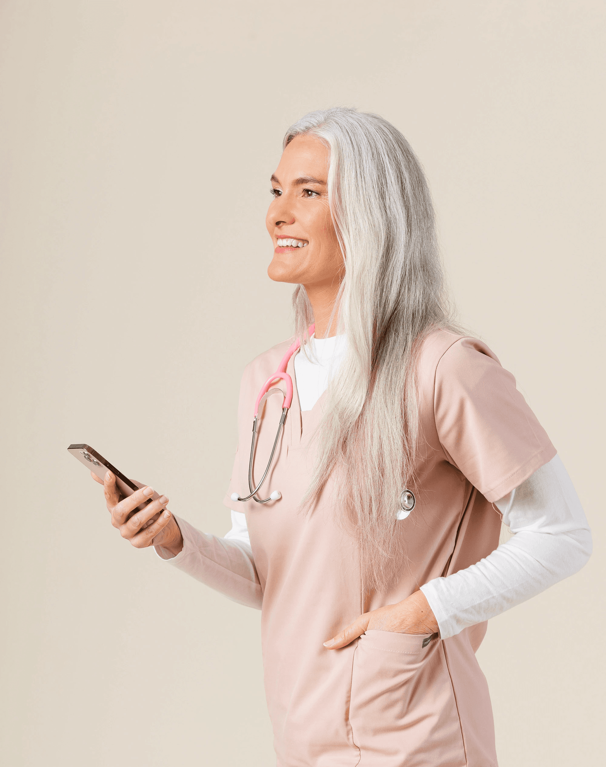 Woman with long gray hair wearing a pink medical uniform, holding a mobile phone and smiling while looking away. A stethoscope hangs around her neck.