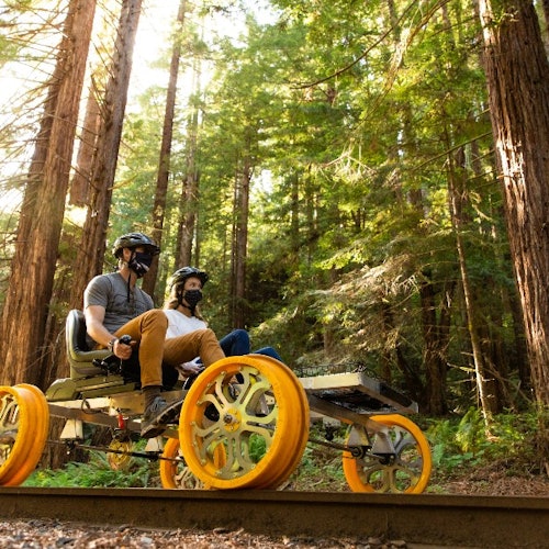 两个人骑着一辆黄色四轮车,沿着轨道穿过阳光照耀的茂密森林。两人都戴着头盔和口罩。