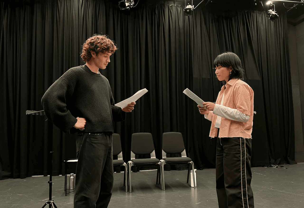A group of people sit around a long table in a brightly lit rehearsal studio, reading and discussing papers in front of them. The room features black curtains, wooden storage cabinets, and large glass doors that look out onto a waterfront boardwalk and industrial steel structures outside. Overhead strip lights and exposed rigging emphasise the professional training environment.