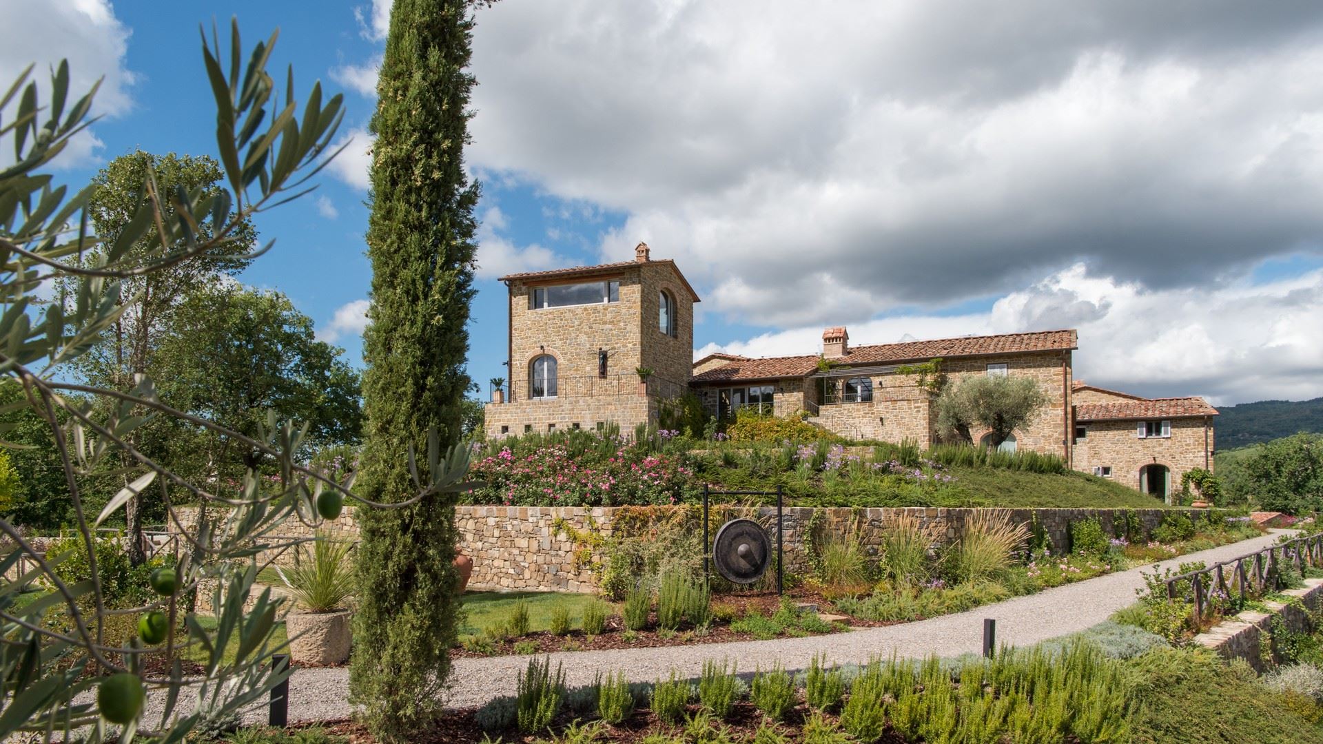 Ein luxuriöses Steinhaus mit Zypressen, Olivenbäumen und Garten im toskanischen Stil unter blauem Himmel.