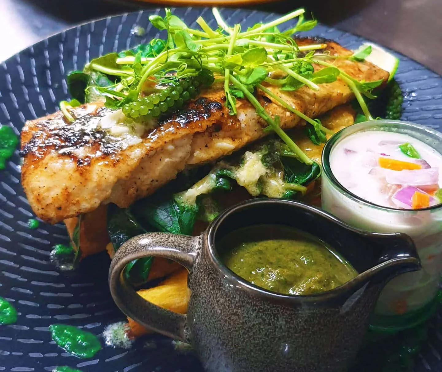 A plate of grilled fish topped with sea grapes and sprouts, served with greens, fries, green sauce, and kokoda.